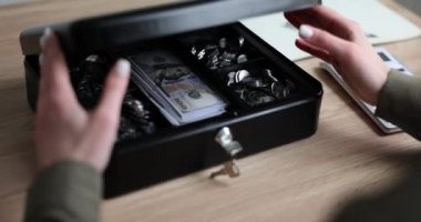 Kadın, muhasebeci kasasını güvenlik kilidi ve dolar banknotlarıyla dolu bir anahtarla açıyor. Üzerinde banknotlar ve madeni paralar olan kara kutu.