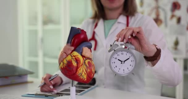 女性医師は ライト病院のオフィスで科学的な心臓モデルの近くに目覚まし時計を保持しています 人間の器官や寿命の概念の詳細な解剖学スローモーション — ストック動画