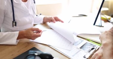 Steteskoplu aile doktoru erkek hasta tedavi planını açıklıyor ve soruları cevaplıyor. Kadın formu doldurur ve erkek ağır çekimde konuşur.