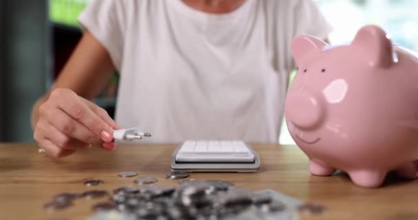 白いシャツの女性は 貯金箱のピンクの切り抜きに電気プラグを挿入しようとします 女性はコインや計算機の近くのテーブルの上に貯金を充電貯金を補充します — ストック動画