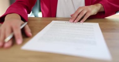 Elinde kalem olan kadın patron, ahşap masada otururken imzalaması için müşteriye sözleşme kağıtlarını veriyor. Pembe ceketli kadın ağır çekimde ticaret anlaşması yapıyor.