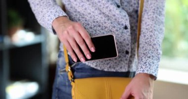 Kadın cihaz kaybını önlemek için akıllı telefonu sarı çantaya koyar. Bluzlu kadın çalışan eşyalarını toplayıp ağır çekimde eve gider.