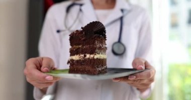 Kadın doktor elinde çikolatalı pasta parçasıyla tabağı tutuyor. Steteskoplu beyaz laboratuar önlüklü kadın klinikteki sağlıksız yiyecekleri parlak pencerelere karşı gösteriyor.