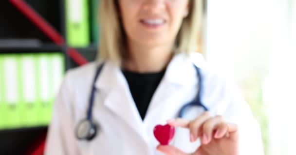 聴診器で笑顔の看護師は小さな赤いハートのおもちゃの姿を与えます 白衣の女性は日当たりの良い診療所のオフィススローモーションに立つ — ストック動画