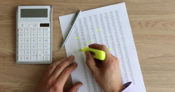 男性の手は 黄色のマーカーペンを使用して財務報告書の重要な数字を強調表示します 会計士は計算機とボールペンで木製の机に座って利益を計算します — ストック動画