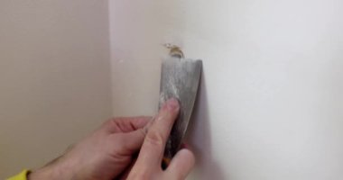 Erkek işçi, sökülmüş vidaların delikleriyle beyaz duvar yüzeyini öğütüyor. İnşa paneli macun bıçağıyla işliyor. Evdeki tamirat işleri.