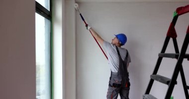 Miğferli ve tulumlu genç bir erkek işçi, duvarları tekerlekli fırçayla boyuyor. Profesyonel müteahhit, odanın iç tarafını yavaş çekimde yeniliyor