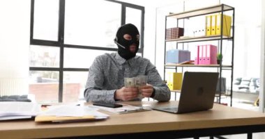 Hırsız maskeli heyecanlı adam ofiste bilgisayarla masada oturan kulaklıklı mikrofona konuşur. Duygusal hacker sinirli omuz silkerek para sayar