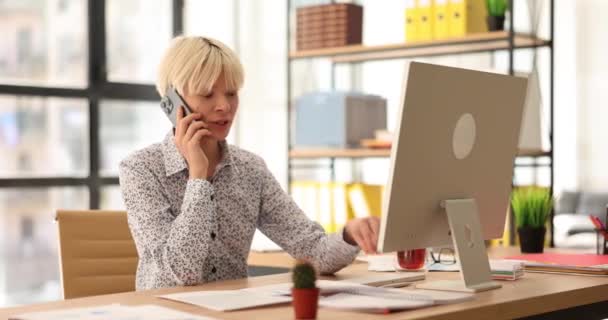 若いビジネスマンの女性がオフィスの机に座って電話で話して笑顔 携帯電話を保持する専門秘書とクライアントとのコンサルティング — ストック動画