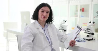 Erkek laboratuvar asistanı onaylıyor. Biyoloji öğrencisi elinde notlarla gülümsüyor ve pano tutuyor.