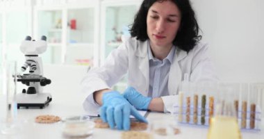 Genç bilim adamı, karabuğday taneli Petri kabını masaya oturtuyor. Lastik eldivenli uzun saçlı adam laboratuvarda yavaş çekimde mahsulleri inceliyor.