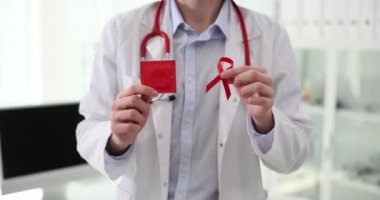Beyaz önlüklü doktor kırmızı kurdele ve prezervatifi elinde tutuyor. AIDS ve HIV farkındalığının uluslararası sembolü ve sağlık korumasının yavaş çekim