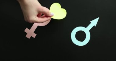 Siyah arka planda pembe kadın ve mavi erkek sembollerinin arasına sarı kağıt kalp koyan bir kadın eli. Kadın ve erkek arasındaki sevgi