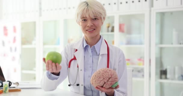 微笑的家庭医生手里拿着青苹果和现实的大脑模型 女专家轮流拿起坐在办公室里慢动作的东西 — 图库视频影像