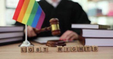 Tahta tokmakla vuran kadına karşı yapılan LGBT hakları cümlesi. Yargıç, gökkuşağı bayrağı ve el yazmaları koleksiyonuyla masada oturarak mahkeme oturumu yürütüyor.