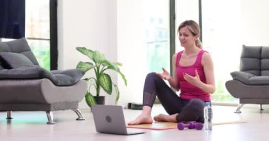 Bayan antrenör, video konferansında öğrencilere ders veriyor. Spor kıyafetli sarışın kadın yoga minderinde oturur ve evdeki dizüstü bilgisayara bakar.