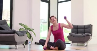 Kadın halterlerle egzersiz yapıyor. Göğsünde şiddetli ağrı hissediyor. Sarışın kadın ellerini göğsüne koyar ve ağır çekimde oturur.