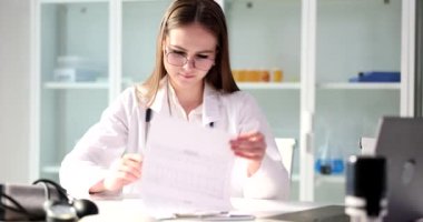 Gülümseyen kadın doktor kağıtlara bakar ve hastanın test sonuçlarını imzalar. İş üniformalı genç stajyer klinik dolaplarına karşı masada oturuyor.