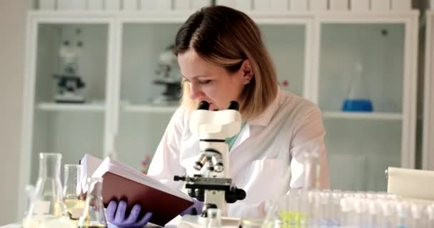Γυναίκα Χημικός Διαβάζει Οδηγίες Ενώ Κοιτάζοντας Μέσα Από Μικροσκόπιο Κάθεται — Αρχείο Βίντεο