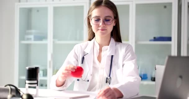 ポジティブな女医者はクリニックのオフィスに座って手に赤いハートのおもちゃを示しています 女性ブルネット人とともにStethoscope少し笑顔遅い動き — ストック動画