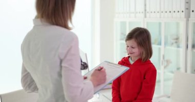 Dosyasıyla gezen kadın doktor hafif okuldaki gülümseyen hastayı muayene ediyor. Okul çocukları için modern yıllık sağlık hizmeti yavaş çekim