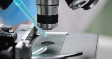 Bilimadamı laboratuvarda mavi reaktifin modern mikroskop altında düşüşünü inceliyor. Lastik eldivenler uzmanı örnek üzerinde detaylı bilimsel araştırmalar yapar.