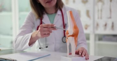 Kadın doktor, modern bir klinikte oturan iskeletin üzerindeki diz çalışmasını açıklıyor. Steteskop ile tıp alanında uzman yavaş çekimde eklemlerin çalışması hakkında konuşuyor