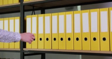 Kadın eli ofis arşivinden sarı yüzük klasörünü alıyor. Modern ofis ağır çekimde geniş rafta organize dizin yapısı ve arşivi