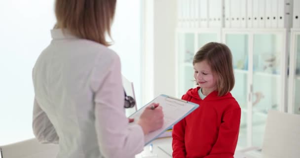クリップボードを持つ女性医師は ライトスクール前提で笑顔の女の子患者を調べます 子供のための現代の年間医療スローモーション — ストック動画