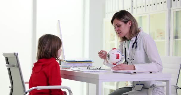 女性医師は 診療所のテーブルに座っているプラスチックモデルの少女患者の目の構造について説明します 女の子慎重に耳を傾けセッションとともに眼科医遅い動き — ストック動画