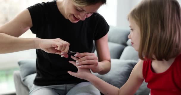 妈妈把坐在轻便公寓沙发上的小女儿的指甲剪掉了 具有镇定表情的学龄前女孩等待着慢动作的结果 — 图库视频影像