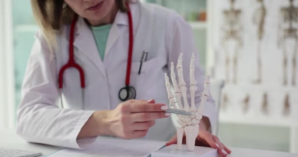 制服を着た女医師が診療所のテーブルに座りスケルトンの手にペン 医学の分野の専門家は腕の部分のスローモーションのスケルトンを提示します — ストック動画