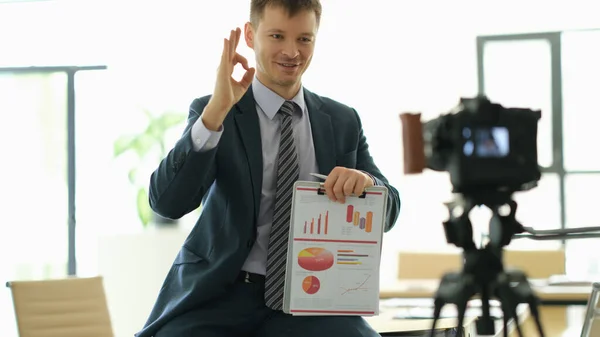 Business coach man showing ok gesture and holding papers with charts in front of camera. Online video conferencing concept