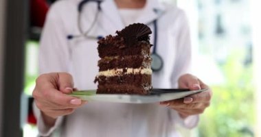 Kadın doktor elinde lezzetli çikolatalı kekle açık pencereye karşı tabağı tutuyor. Üniformalı kadın klinikte tatlı gösteriyor. Sağlıksız beslenme