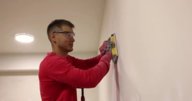 Pozitif işçi ışık odasında zımpara makinesiyle alçıpan parlatıyor. Gözlüklü inşaatçı yenilenmiş muhafazayı yerleştirdikten sonra yüzeyi öğütüyor