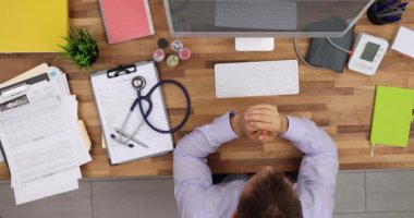 Doktor hasta bilgileriyle birlikte steteskop yakınındaki tahta masada oturuyor. Adam bilgisayar ekranına yavaş çekimde bakmayı düşünüyor.