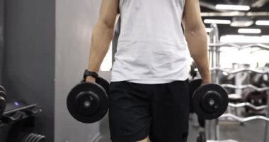 Güçlü adam elinde ağır dambıllarla salonda yürüyor. Spor kıyafetlerindeki formda sporcu ağır çekimde yoğun eğitime hazırlanıyor.