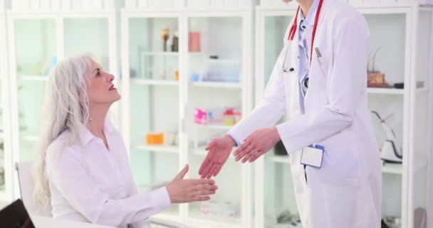 ブロンドの医者はクリニックのオフィスで成熟した女性クライアントの手を振る 感情的な女性の笑顔を迎える フレンドリーな挨拶と患者治療のコンセプトスローモーション — ストック動画