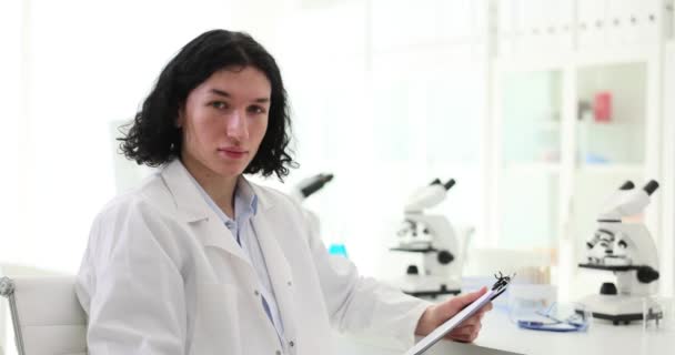 拥有长发的男性科学家拿着剪贴板坐在桌旁 带着显微镜 年轻的化学家在配备现代化设备的实验室工作 — 图库视频影像