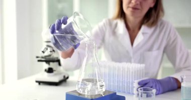 Lastik eldivenli kadın laboratuvar teknisyeni cam şişeye temiz su döküyor. Profesyonel bilim adamı çözümleri laboratuvar karıştırıcısı kullanarak karıştırıyor