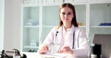 Genç bayan doktor kameraya bakarken gözlük takıyor. Tıbbi üniformalı ciddi bir kadın klinikte dizüstü bilgisayarın yanında ağır çekimde oturuyor.