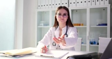 Gözlüklü genç bayan doktor elini uzatıp yardım teklif ediyor ve oturmak istiyor. Üniformalı kadın dizüstü bilgisayarın yanında oturuyor ve ağır çekimde kameraya bakıyor.