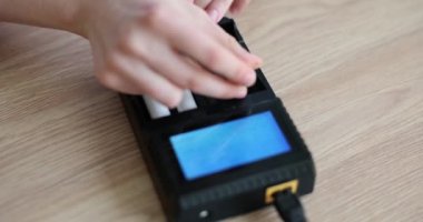 Kadın batarya için hücreleri tahta masadaki şarj cihazına yerleştiriyor. Güç elementi yeniden yükleniyor. Kadın ağır çekimde elektronik alet kullanıyor.