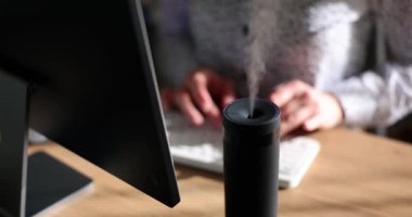 Bilgisayarın klavyesinde aroma yayıcı nemlendirici ile oturan kadın yönetici tipleri. Ofis çalışanı, iş yerinde ağır çekimde gerekli yağların kokusundan hoşlanır.