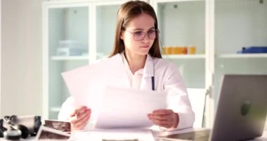 Gözlüklü kadın doktor klinikte dizüstü bilgisayarın yanında oturan belgeleri kontrol ediyor. Tıbbi çevredeki genç kadın evrak işleriyle meşgul.
