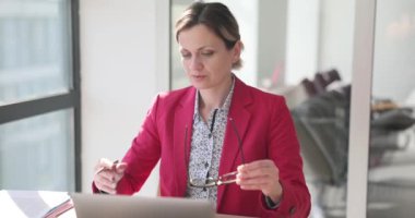 Pembe ceketli bayan yönetici gözlüğünü takıp ofiste dizüstü bilgisayarda çalışmaya hazırlanıyor. Odaklanmış kadın, web sitesindeki proje istatistiklerini yavaş çekimde kontrol ediyor