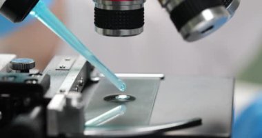 Kimyager, modern mikroskop altında donanımlı laboratuvarda mavi reaktifin düşüşünü inceliyor. Lastik eldivenli bilim adamı sıvı üzerinde detaylı bilimsel araştırma yapıyor.