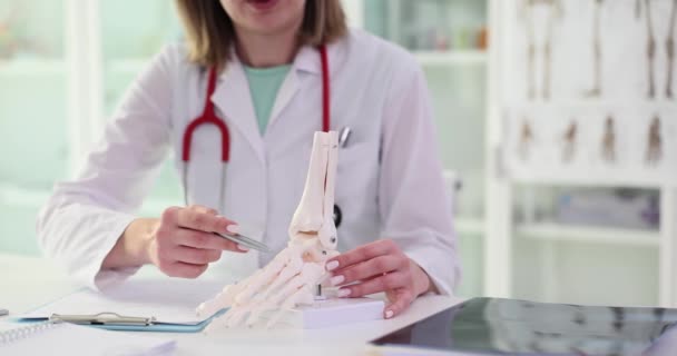 女性医師は 軽い病院の前提でテーブルに座って足の骨格の人工モデルに骨にペンを指す 女性は人間の関節の構造を示すスローモーション — ストック動画