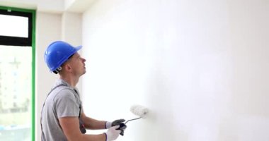 Erkek işçi, düz ışık odasının beyaz tamir odasındaki duvarı boyuyor. Miğferli ressam yüzeyde beyaz renk ve silindir kullanır. Ev içi gelişim yavaş çekim