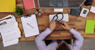 Erkek doktor bilgisayarda çalışırken steteskop takıyor. Hastalarla konsültasyonlar hakkında rapor dolduruyorum. Uzman, ahşap masada oturuyor.